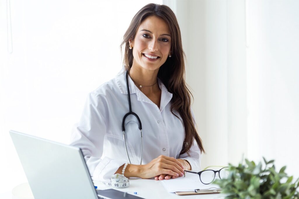 A woman in a white coat sits at a desk with a stethoscope around her neck, smiling as she rests her hands on the desk. Beside her laptop, papers, and a plant in the foreground, an Ace QBank for MCCQE1 preparation is subtly visible.