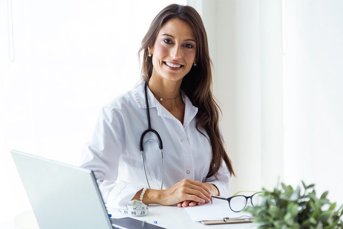 A woman in a white coat sits at a desk with a stethoscope around her neck, smiling as she rests her hands on the desk. Beside her laptop, papers, and a plant in the foreground, an Ace QBank for MCCQE1 preparation is subtly visible.