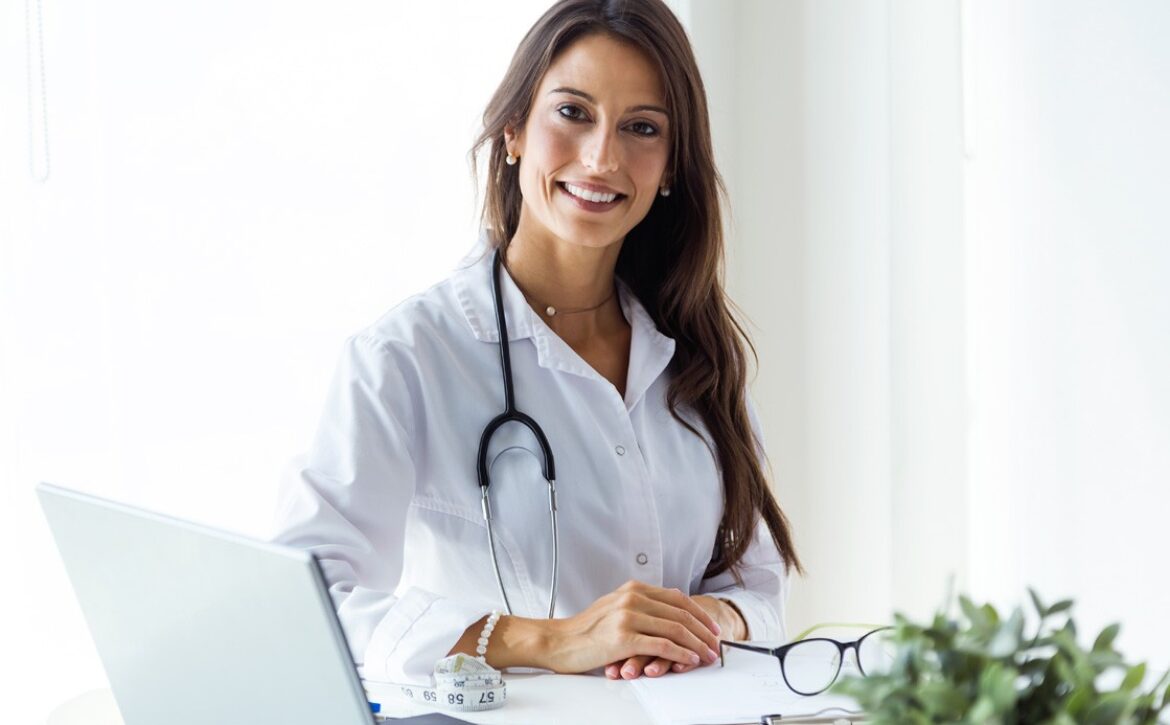 A woman in a white coat sits at a desk with a stethoscope around her neck, smiling as she rests her hands on the desk. Beside her laptop, papers, and a plant in the foreground, an Ace QBank for MCCQE1 preparation is subtly visible.