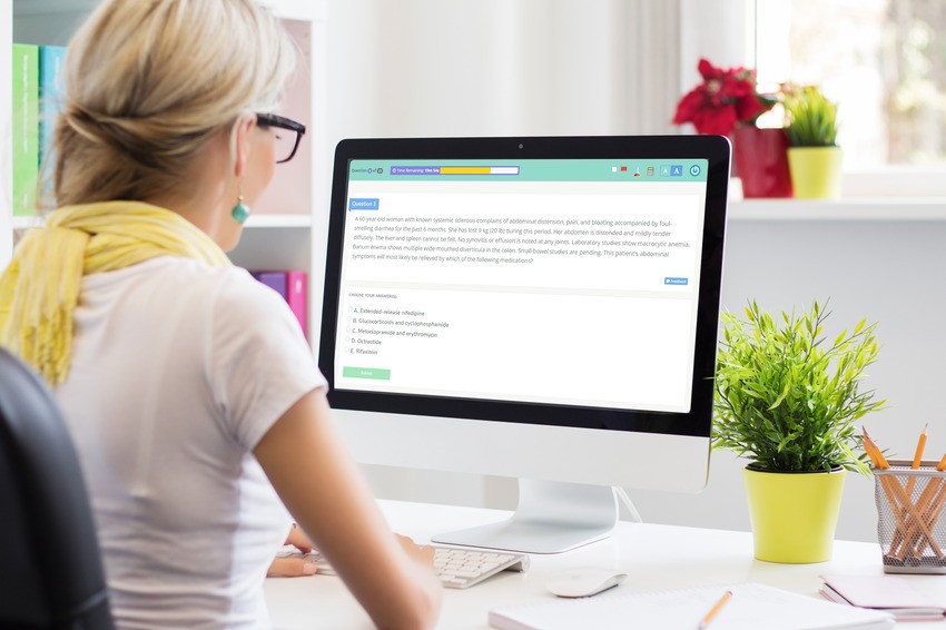 A person wearing glasses and a white shirt is sitting at a desk, looking at a computer screen displaying an MCCQE1 exam from Ace QBank. The desk has a potted plant and some colorful books nearby.