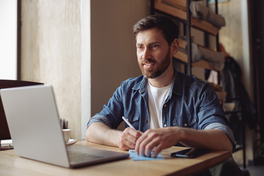 A man in casual clothing sits at a wooden table with a laptop, taking notes on a notepad with a pen, and smiling while looking at the screen. He's immersed in his MCCQE1 exam preparation using Ace QBank.