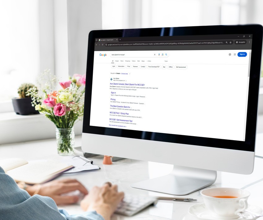 Person typing on a keyboard at a desk with a computer showing a Google search results page for MCCQE1 exam preparation. A vase with flowers and a cup on a saucer are also on the desk, alongside an open Ace QBank, suggesting quick shortcuts for MCCQE1 success.