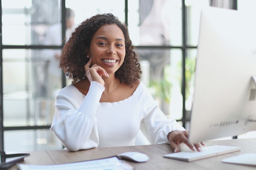 A woman in a white blouse is sitting at a desk, smiling, and working on her computer, utilizing Ace QBank for her MCCQE1 exam preparation.