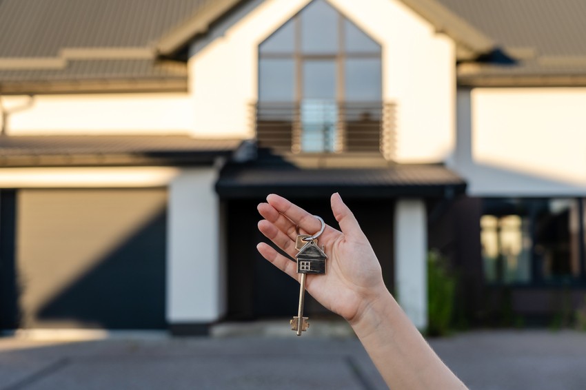 Close-up of a hand holding a key with a house-shaped keychain in front of a modern house, symbolizing how Acing MCCQE1 can unlock your future in Canada.