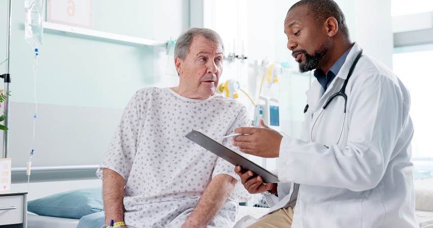 A doctor in a white coat has passed the MCCQE1 exam talking with an elderly male patient seated on a hospital bed. Medical equipment can be seen in the background, highlighting the Crucial Role of Tutor & Time Mode in MCCQE1 exam prep.