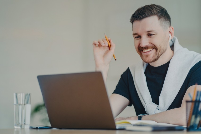 A person wearing a sweatshirt smiles while using a laptop at a desk. They are holding a pencil with a notebook and a glass of water nearby, preparing to ace the MCCQE1 with Ace QBank and unlock their future in Canada.