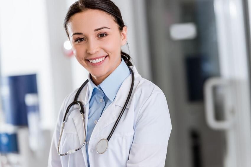 A woman in a white lab coat and stethoscope smiles confidently while standing in a medical setting, embodying the success that comes from diligent MCCQE1 exam preparation with resources like Ace QBank.