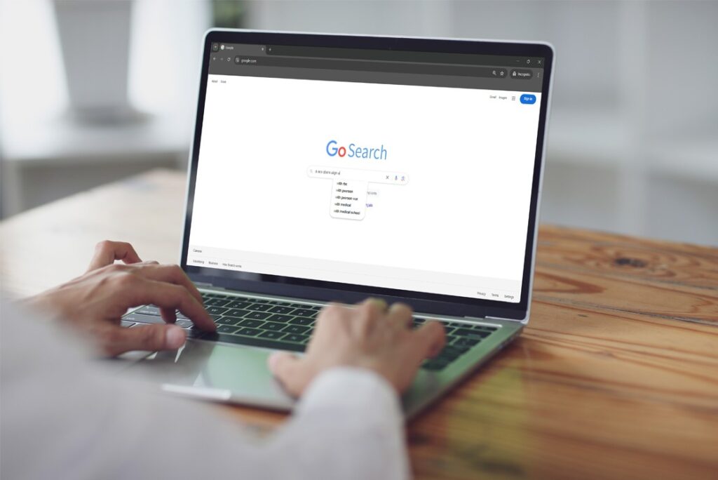 Person typing on a laptop with a search engine page open displaying "GoSearch" and a drop-down list of search suggestions, including "MCCQE1 exam preparation." The laptop sits on a wooden table.