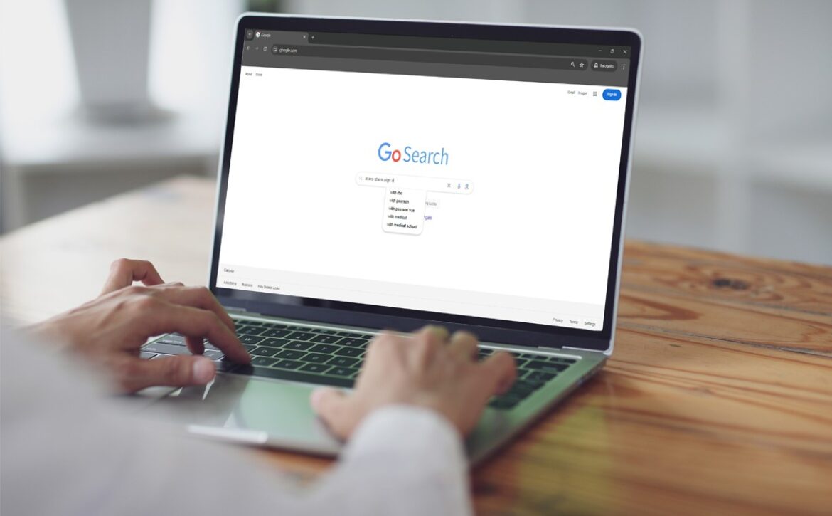 Person typing on a laptop with a search engine page open displaying "GoSearch" and a drop-down list of search suggestions, including "MCCQE1 exam preparation." The laptop sits on a wooden table.