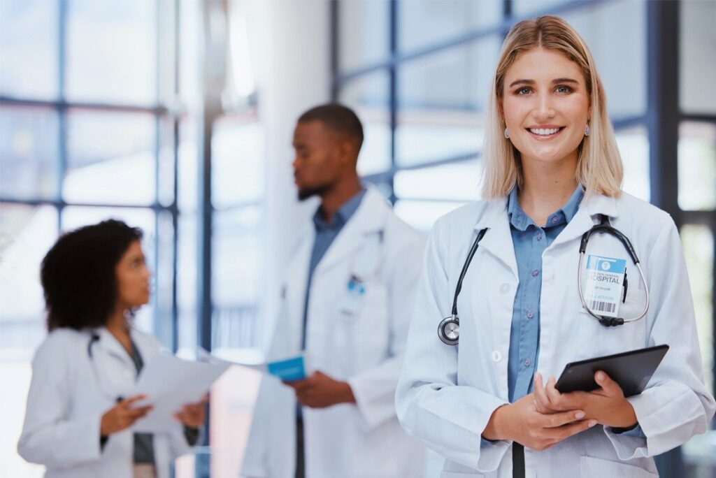 A smiling woman in a white coat holding a tablet stands in the foreground, with two other doctors in white coats discussing the Canadian Medical Licensing Exam pass rate in the background.