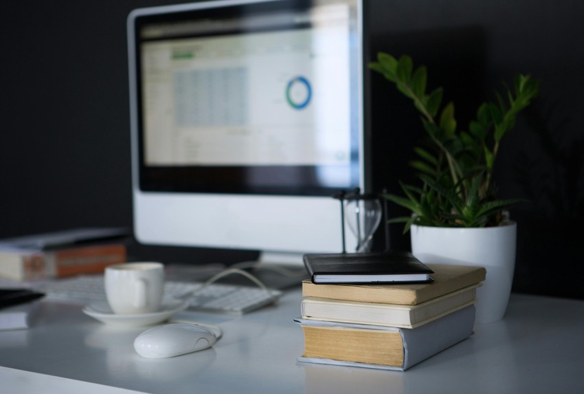 A workspace with a computer monitor displaying a chart, books on Ace QBank for MCCQE1 exam preparation, a notebook, a white computer mouse, a cup, and a potted plant on a white desk.