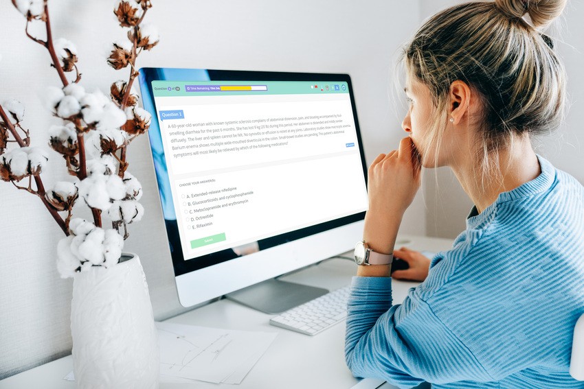 A person in a blue striped shirt is sitting at a desk, looking at a computer screen displaying MCCQE1 exam preparation material. A vase with cotton stems is on the desk next to the monitor.