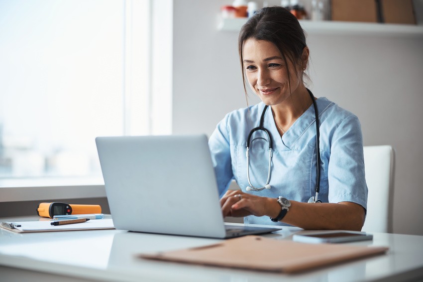 A nurse in blue scrubs sits at a desk, smiling and typing on a laptop, likely reviewing her Ace QBank scores. A stethoscope is around her neck, with a clipboard and other items on the desk, as she prepares for the MCCQE1.