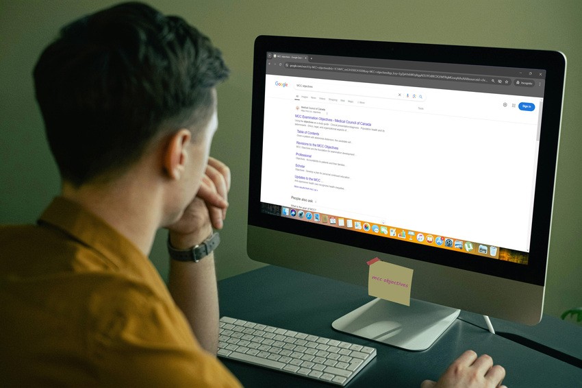 A person is sitting at a desk and a sticky note labeled "MCC objectives" is attached to the monitor. They're clearly deep into MCCQE1 exam preparation, possibly searching for resources like Ace QBank.