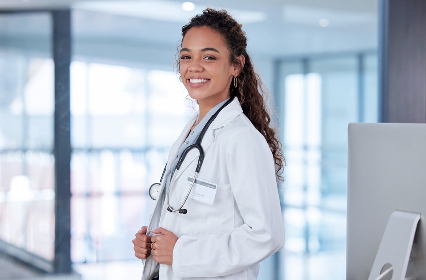 A woman wearing a white lab coat and stethoscope, stands in a medical facility with glass windows in the background, Studying for MCCQE1 exam preparation using Ace QBank.