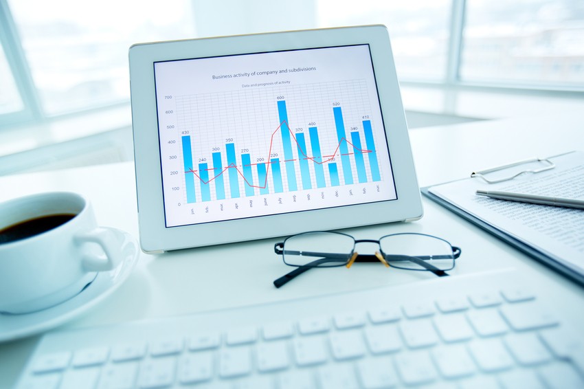 A tablet displaying a business activity graph sits on a desk with a cup of coffee, eyeglasses, a keyboard, and a document showing the MCCQE Part 1 pass rate.