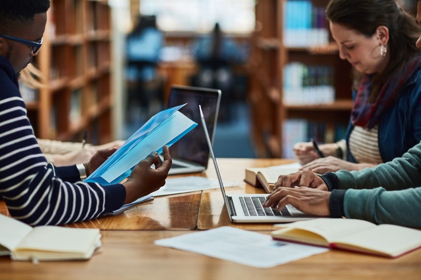 People sitting at a library table, using laptops and reading documents, likely preparing for the MCCQE1. Bookshelves filled with resources are visible in the background.