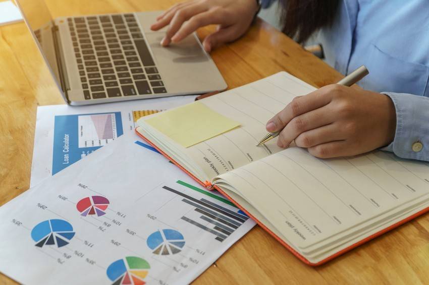 A person is working on a laptop with a notebook in hand, reviewing various documents with pie charts and graphs spread on the wooden table. Amid the busy scene, a tab open to Ace QBank suggests preparation for an upcoming MCCQE1 exam.