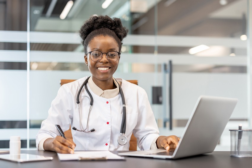 A doctor wearing a white coat with a stethoscope around the neck is seated at a desk, smiling, and writing on a document with a laptop open in front while studying for MCCQE1 using Ace QBank.