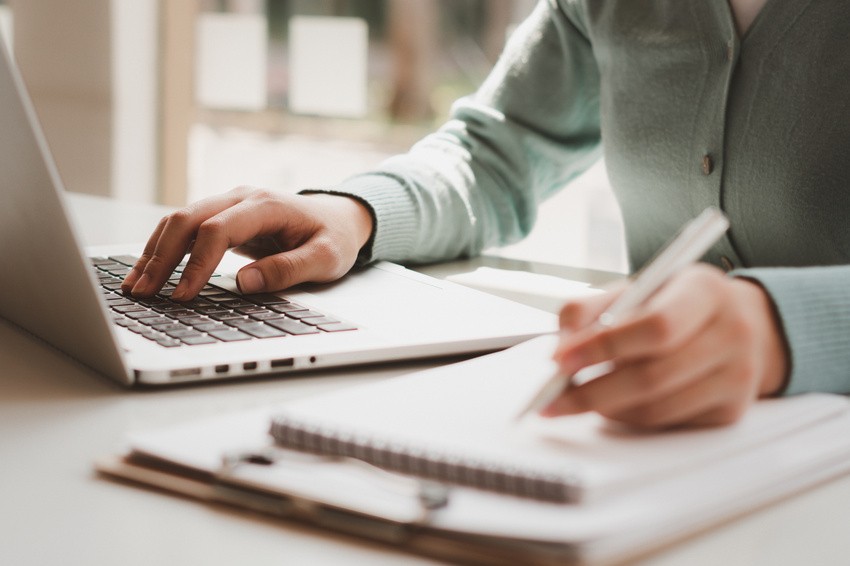 A person is diligently typing on a laptop while jotting down notes in a notebook at the table, possibly preparing for the MCCQE1 with the reliable Ace QBank as their study companion.