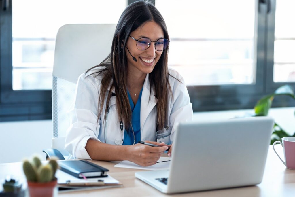 A woman wearing glasses and a white coat is sitting at a desk, smiling while using her laptop with Ace QBank for MCCQE1 prep. She has a headset on and holds a pen, embodying the future of telemedicine.