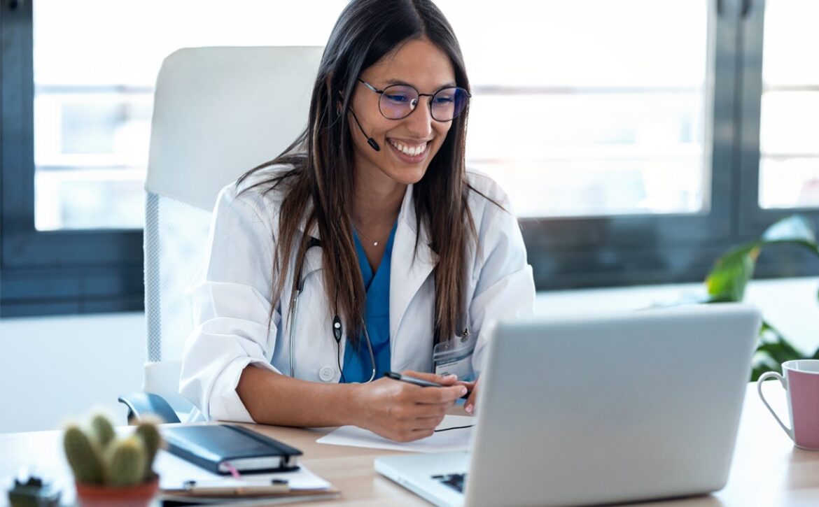 A woman wearing glasses and a white coat is sitting at a desk, smiling while using her laptop with Ace QBank for MCCQE1 prep. She has a headset on and holds a pen, embodying the future of telemedicine.