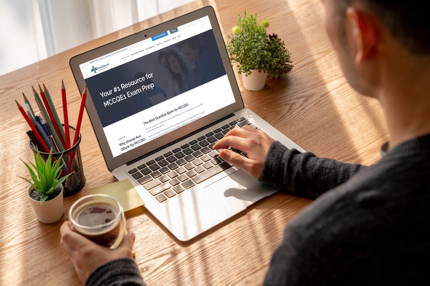 A person uses a laptop displaying a webpage titled "MCCQE part 1 Exam Prep" alongside the latest MCCQE1 resources. They hold a cup, while the table features a plant and a container of colored pencils, adding to an inspiring study atmosphere.