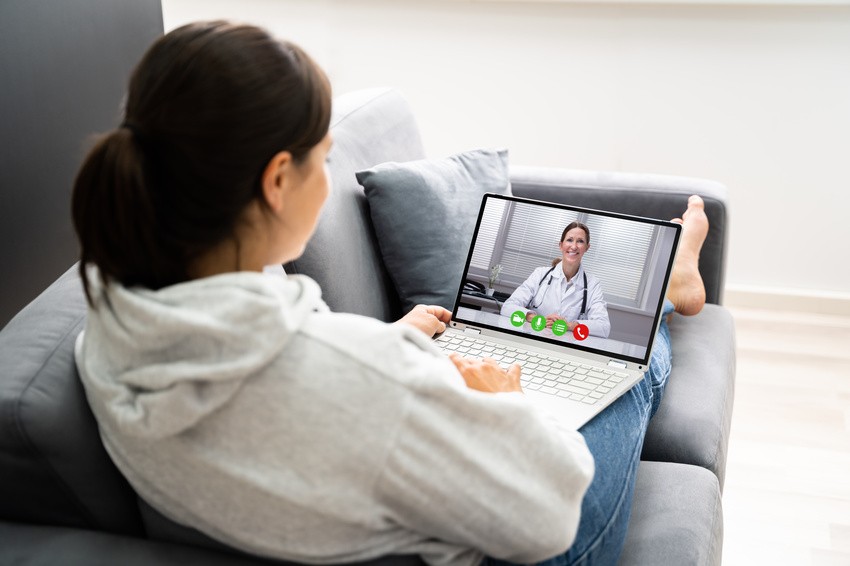 A person in a hoodie sits on a couch, using a laptop for a video call with a doctor, possibly discussing strategies from Ace QBank to ace the MCCQE1 exam or is telemedicine.