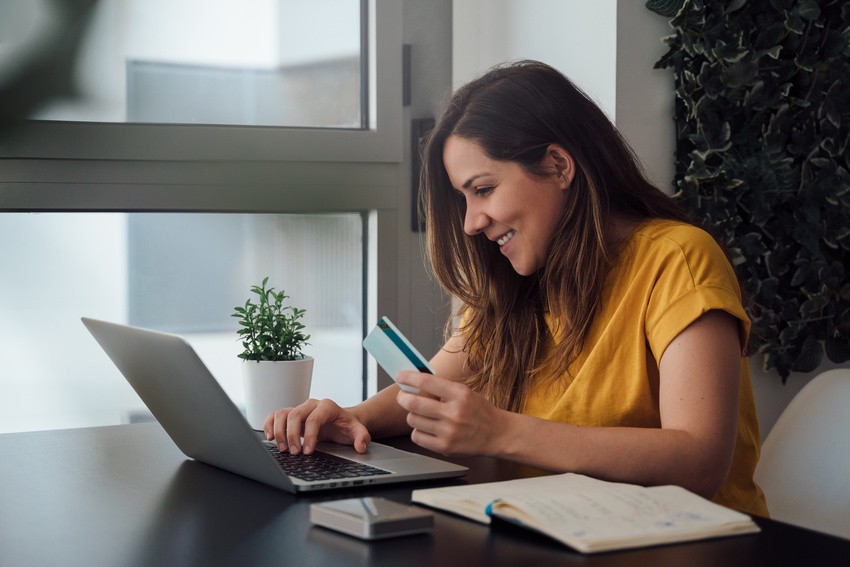 A woman is sitting at a table, holding a credit card and typing on her laptop, exploring the Ace QBank Update for MCCQE1. A small plant is beside her, and she considers trying the demo account.