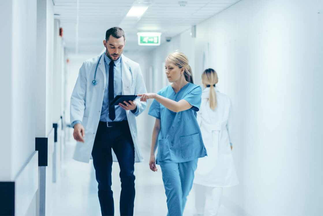 A doctor and a nurse stroll down the hospital corridor, reviewing Canadian guidelines on a tablet. Ahead, another medical professional in a white coat navigates using Ace QBank to prepare for MCCQE1.