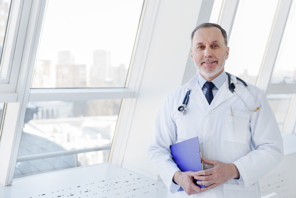 A doctor wearing a white coat and stethoscope stands by large windows, holding a blue folder filled with self-assessment notes from Ace QBank to excel in the MCCQE1.