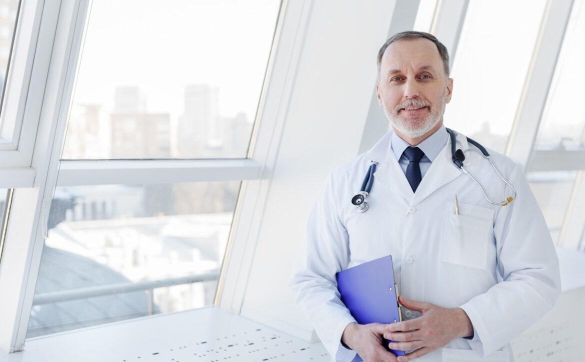 A doctor wearing a white coat and stethoscope stands by large windows, holding a blue folder filled with self-assessment notes from Ace QBank to excel in the MCCQE1.