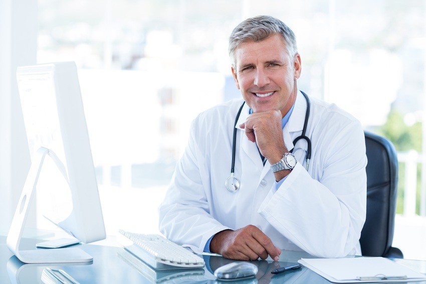 A doctor in a white coat sits at a desk with a computer, smiling confidently. With the latest Ace QBank Update for MCCQE1 on his screen, he's ready to tackle challenges. A stethoscope hangs around his neck, symbolizing his dedication to patient care and medical excellence.