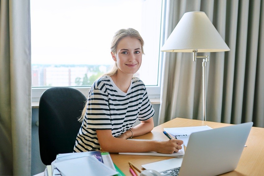 A person in a striped shirt sits at a desk with a laptop, Ace QBank opened beside them, and books piled high. Under the warm glow of the lamp, they smile at the camera, fully focused and prepared on how to study for MCCQE1 distraction-free.