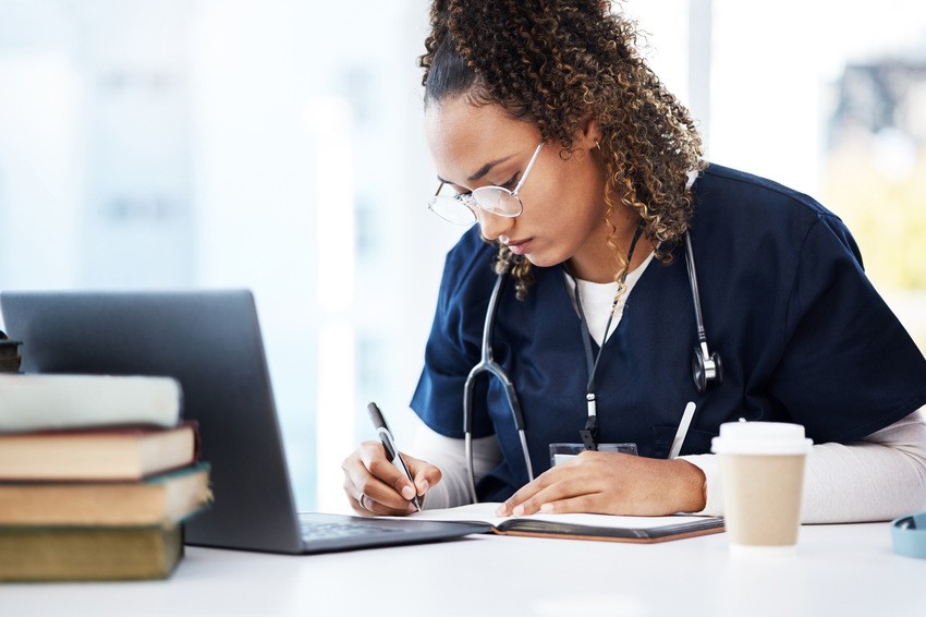 A person in blue scrubs is sitting at a desk, focused on writing in a notebook. Nearby are a laptop open to Ace QBank and stacked books for MCCQE1 preparation. A stethoscope hangs around their neck while a coffee cup keeps them energized for distraction-free study sessions.