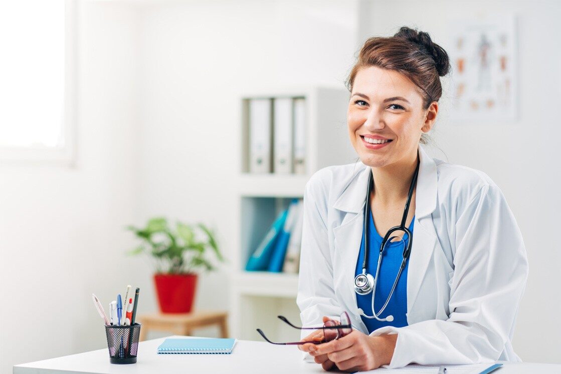 A smiling doctor in a white coat, preparing for the Ace QBank Update for MCCQE1, holds glasses while sitting at a desk adorned with office supplies and a potted plant.