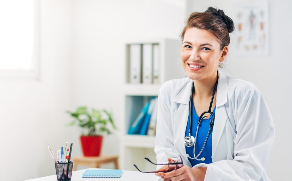 A smiling doctor in a white coat, preparing for the Ace QBank Update for MCCQE1, holds glasses while sitting at a desk adorned with office supplies and a potted plant.