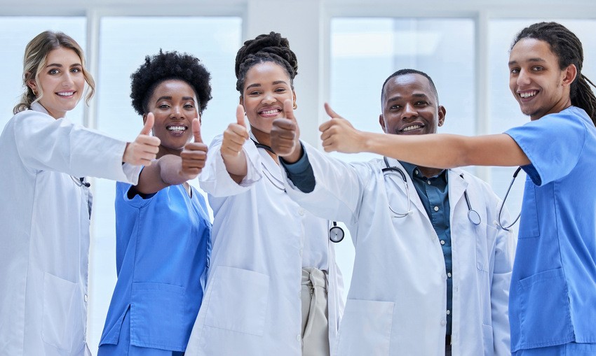 A group of five healthcare professionals in lab coats and scrubs stand indoors, smiling and giving a thumbs-up gesture, celebrating their success with Ace QBank as a tool for MCCQE1 self-assessment.