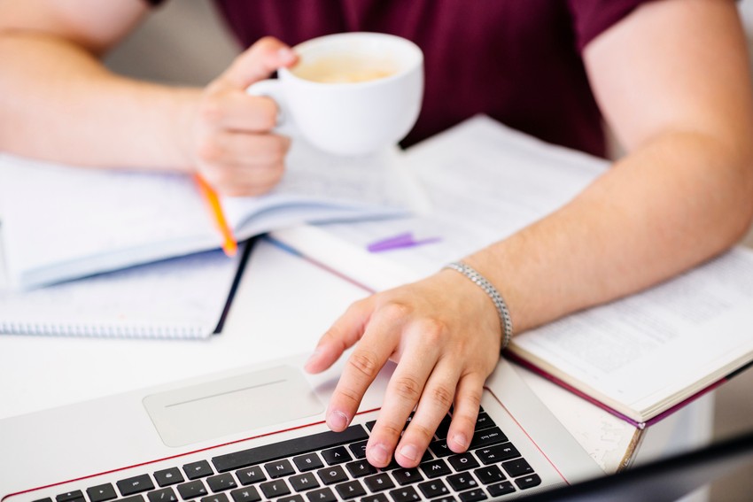 A person holds a cup of coffee while typing on a laptop surrounded by open notebooks and papers, exploring Ace QBank strategies to study for the MCCQE1 distraction-free.