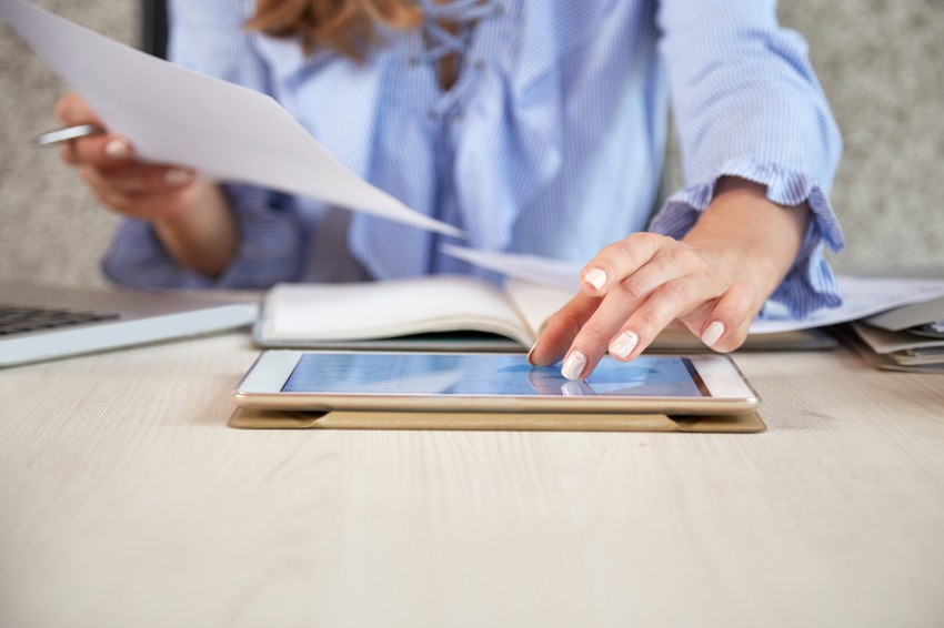 A person in a blue shirt uses a tablet and holds documents, with a laptop and an open book on the table. They efficiently navigate Ace QBank resources and review Canadian guidelines, preparing meticulously for the MCCQE1 exam.