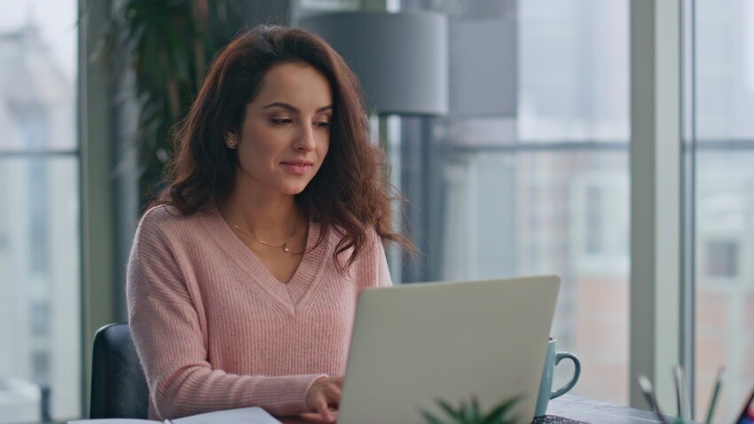 In a bright office with large windows, a woman in a pink sweater is diligently preparing for the MCCQE1, using Ace QBank on her laptop. The desk is organized with a notebook, pen, and mug, creating an ideal distraction-free study environment.