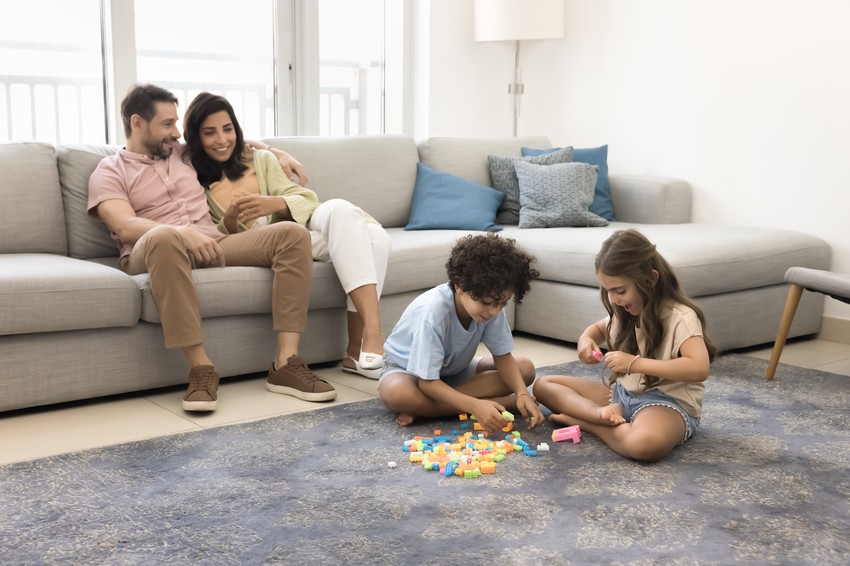 A family sits on a gray sofa, their peaceful living room free from distractions, while two children play with colorful blocks on the carpeted floor. It's an ideal setting for parents to browse guides like "How to Study for MCCQE1 Distraction-Free?"