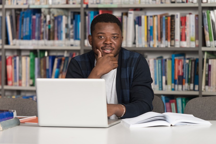 A person sits at a table in a library with a laptop and open book, immersed in mastering the AMC MCQ Exam, surrounded by shelves of books.