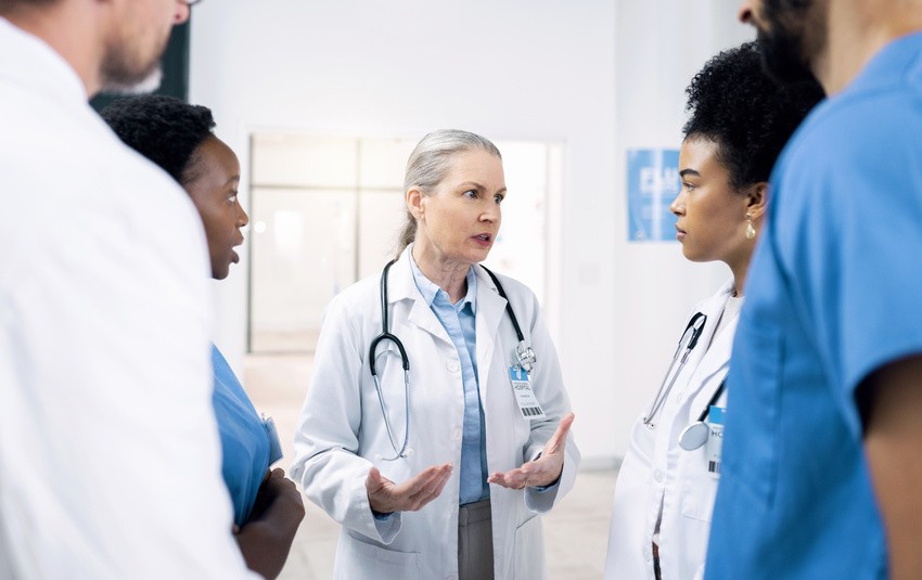 A group of medical professionals in discussion, with a senior doctor explaining the new MCCQE1 format to three others in a hospital setting.