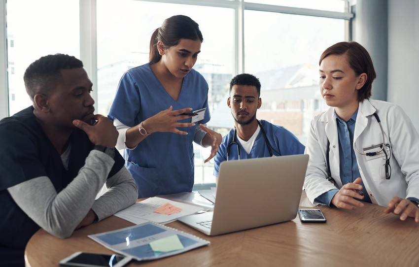 Four medical professionals in scrubs and coats discuss MCCQE1 exam preparation while looking at a laptop in a bright office setting, utilizing Ace Qbank to adapt to the new MCCQE1 format.