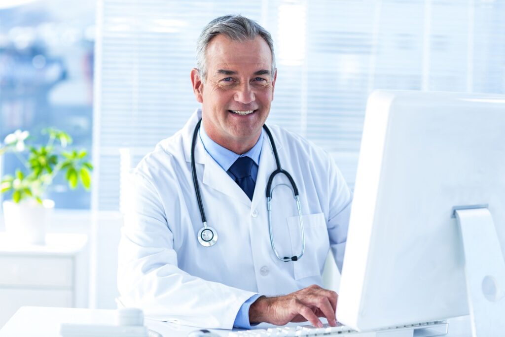 A doctor in a white coat with a stethoscope, seated at a desk, smiling at the camera in a well-lit office setting, confidently discusses strategies for MCCQE1 exam preparation and Ace QBank role.