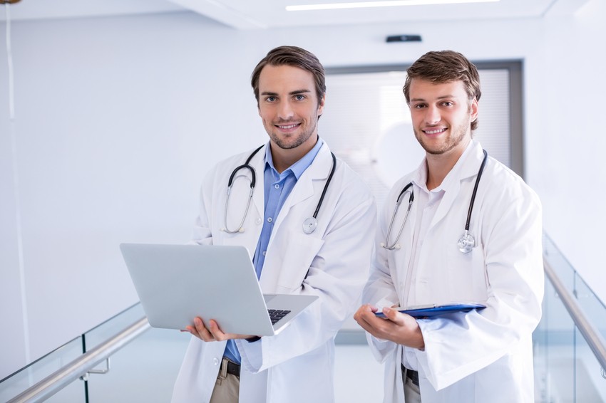 Two doctors in white coats stand in a hallway, embodying the focus and diligence needed for mastering the AMC MCQ Exam. One holds a laptop and the other holds a clipboard, both wearing stethoscopes around their necks as they consult on Ace QBank strategies.