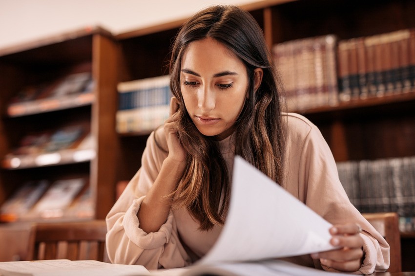 A woman, engrossed in study, flips through pages at a library table surrounded by bookshelves. She's delving into MCC objectives, preparing for the anticipated changes to the MCCQE Part I in 2025.