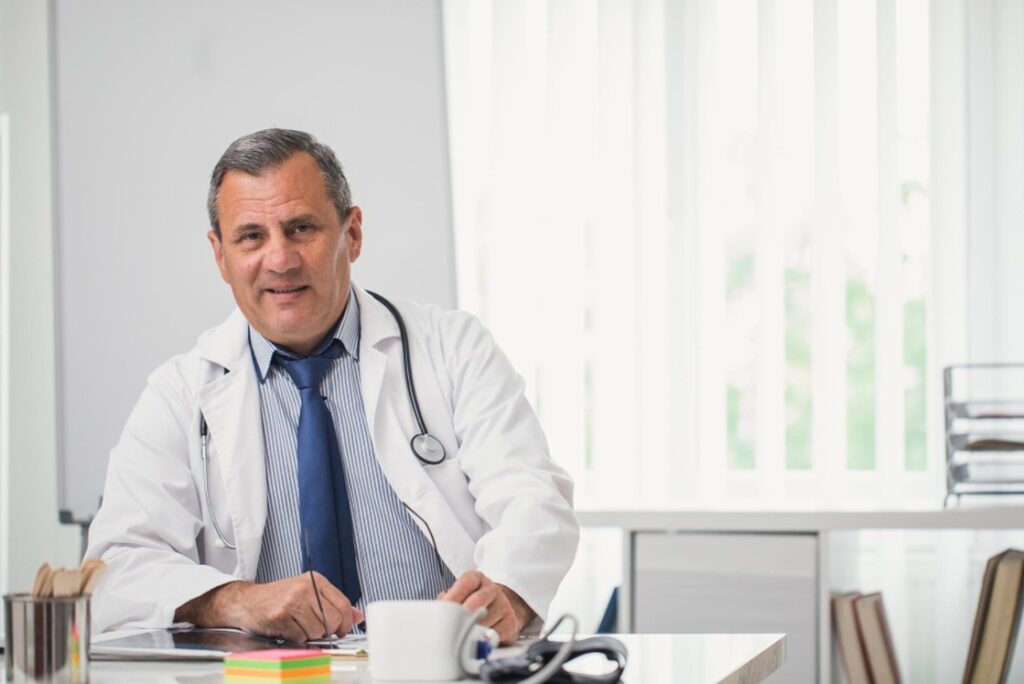 A middle-aged male doctor in a white coat and stethoscope sits at his desk, an Ace QBank open beside him, smiling confidently as he reviews MCCQE1 Exam Dates with office supplies and a mug close by.