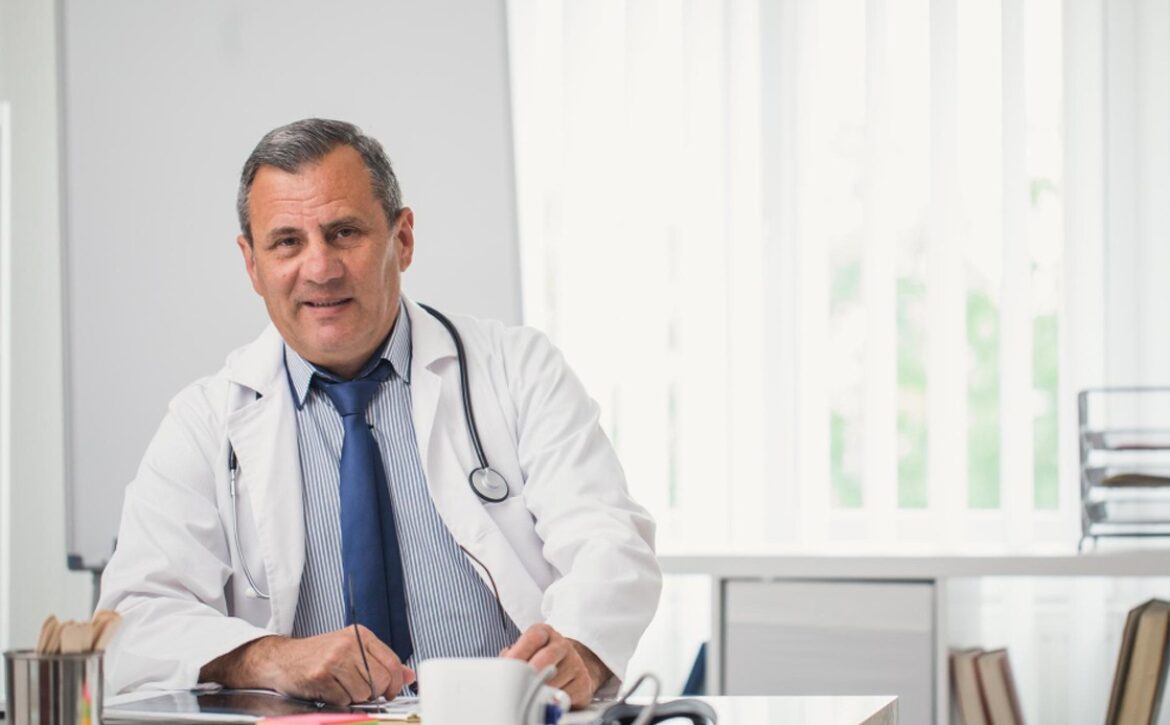 A middle-aged male doctor in a white coat and stethoscope sits at his desk, an Ace QBank open beside him, smiling confidently as he reviews MCCQE1 Exam Dates with office supplies and a mug close by.